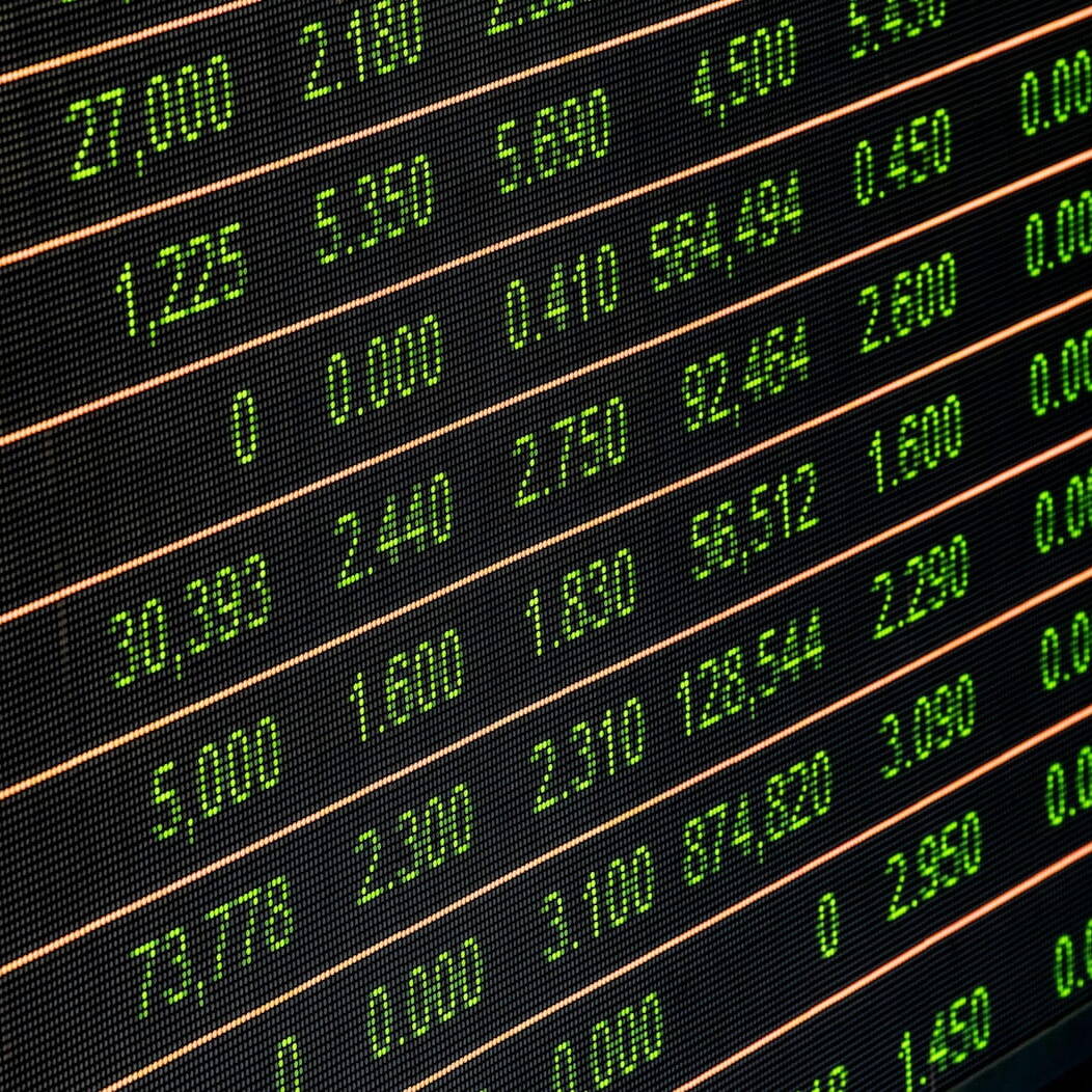Foto en primer plano de una gran pantalla LED de cotizaciones bursátiles que muestra cotizaciones en vivo con números verdes y naranjas, líneas de precios, volúmenes y nombres de empresas en árabe y cifras, ilustrando la actividad dinámica de los mercados financieros globales.
