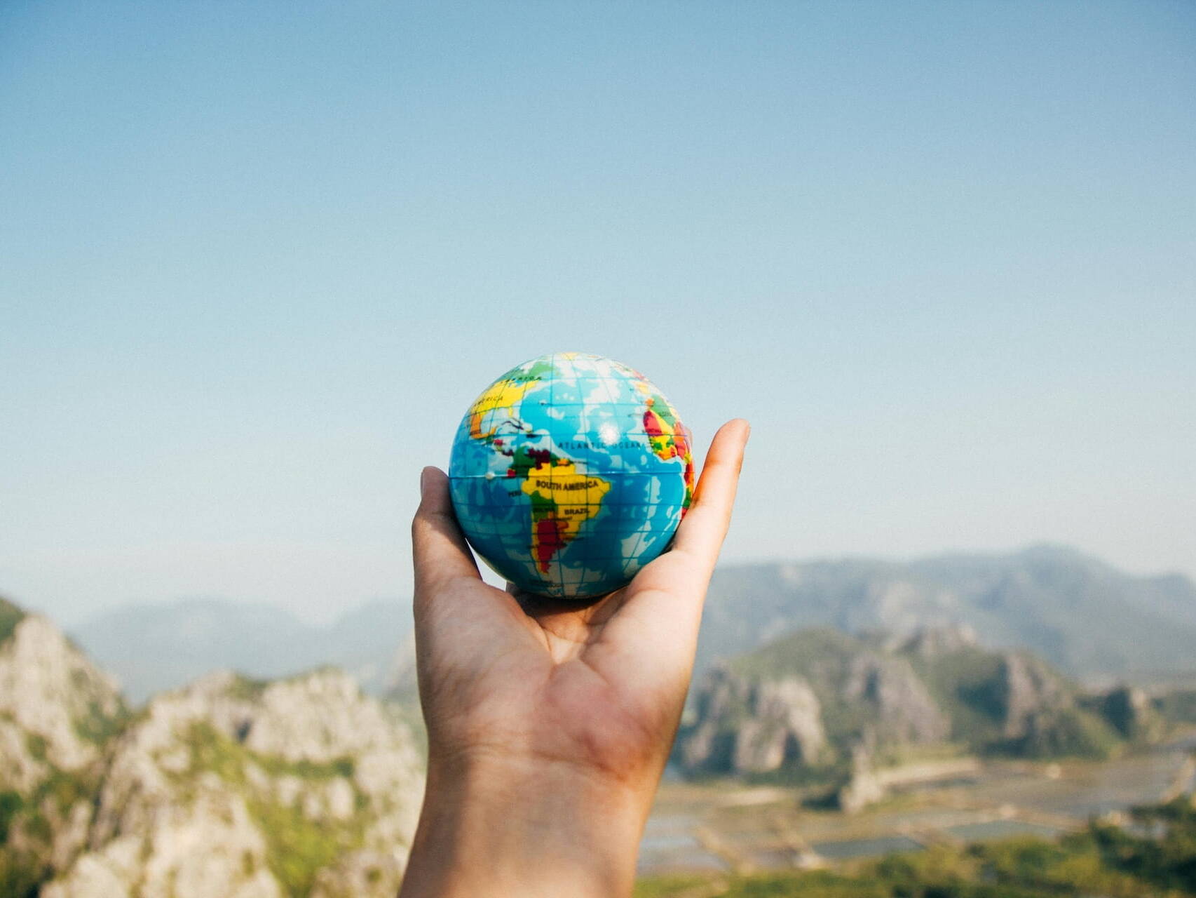 Mano sosteniendo un pequeño globo terráqueo contra un paisaje montañoso, simbolizando la internacionalización del patrimonio familiar, la diversificación global de activos y la gestión estratégica transfronteriza.